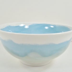 May include: A white ceramic bowl with a light blue interior and a light blue band around the middle. The bowl has a rounded shape and a smooth, glossy finish. Suitable for serving food, it is a decorative item.