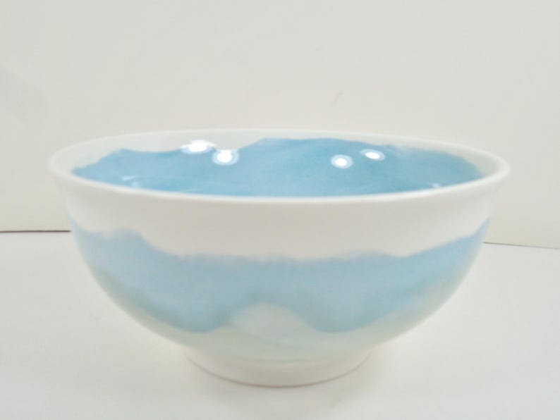 May include: A white ceramic bowl with a light blue interior and a light blue band around the middle. The bowl has a rounded shape and a smooth, glossy finish. Suitable for serving food, it is a decorative item.