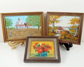 May include: Three framed landscape paintings. One depicts a white house in a field, another a red covered bridge, and the third a vase of orange flowers. The paintings are in ornate gold and brown frames, displayed on decorative stands.