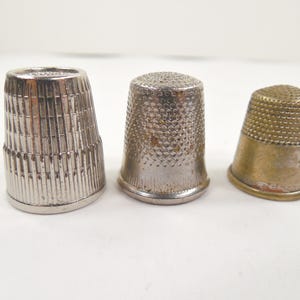 May include: Three metal thimbles of varying designs and colors are displayed. The thimbles are silver and gold in color. The thimbles are used for sewing and are made of metal. The thimbles are of different sizes and shapes.