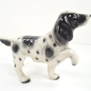 May include: A ceramic figurine of a dog in a playful pose. The dog has a white body with black spots and a black head and ears. One front leg is raised, giving the impression of movement. The figurine is on a white surface.