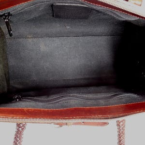 May include: The interior of a brown leather handbag featuring a black fabric lining. The bag includes a central compartment with a zippered pocket on one side and a rectangular leather patch logo centered on the opposite side, with braided handles.