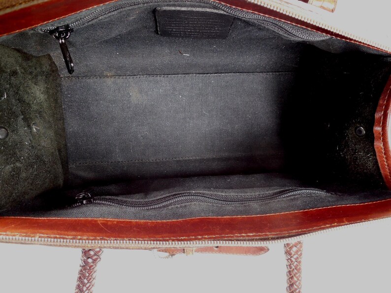 May include: The interior of a brown leather handbag featuring a black fabric lining. The bag includes a central compartment with a zippered pocket on one side and a rectangular leather patch logo centered on the opposite side, with braided handles.