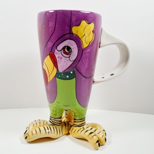 May include: A purple ceramic mug with a cartoon bird design. The mug has a white handle and yellow, green, and red accents. The mug has three yellow and black feet.