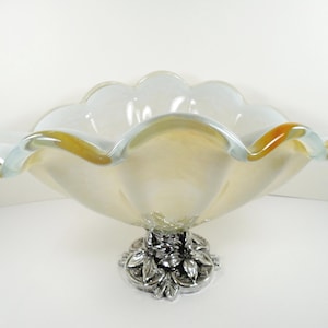 May include: A decorative glass bowl with a ruffled edge and a silver-colored floral base. The bowl has a creamy white interior with a golden-orange rim and a light blue edge. A stylish centerpiece.