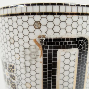 May include: Close-up of a white ceramic mug with a black handle. The mug features a geometric design of black squares and hexagons, with gold and black trim. The mug has a unique, mosaic-like pattern.