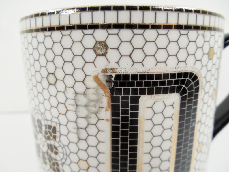 May include: Close-up of a white ceramic mug with a black handle. The mug features a geometric design of black squares and hexagons, with gold and black trim. The mug has a unique, mosaic-like pattern.