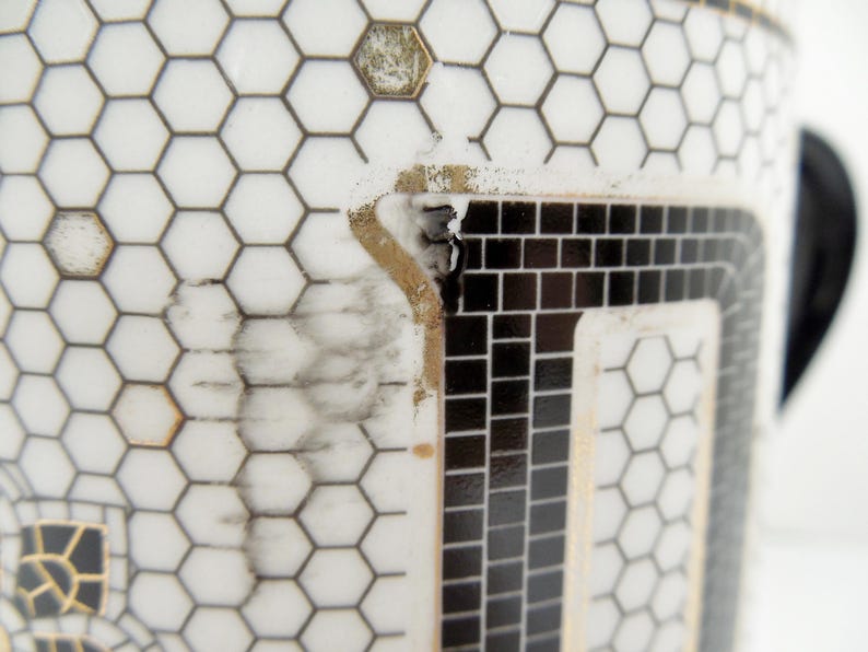 May include: Close-up of a white ceramic mug with a black and gold geometric pattern. The design features a honeycomb pattern and a rectangular mosaic design. The mug has a black handle.