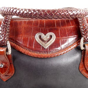 May include: A two-tone handbag featuring a smooth black leather base and a brown crocodile-embossed leather upper section. The bag includes a braided brown leather handle and a central silver-toned metal heart emblem with intricate scrollwork.