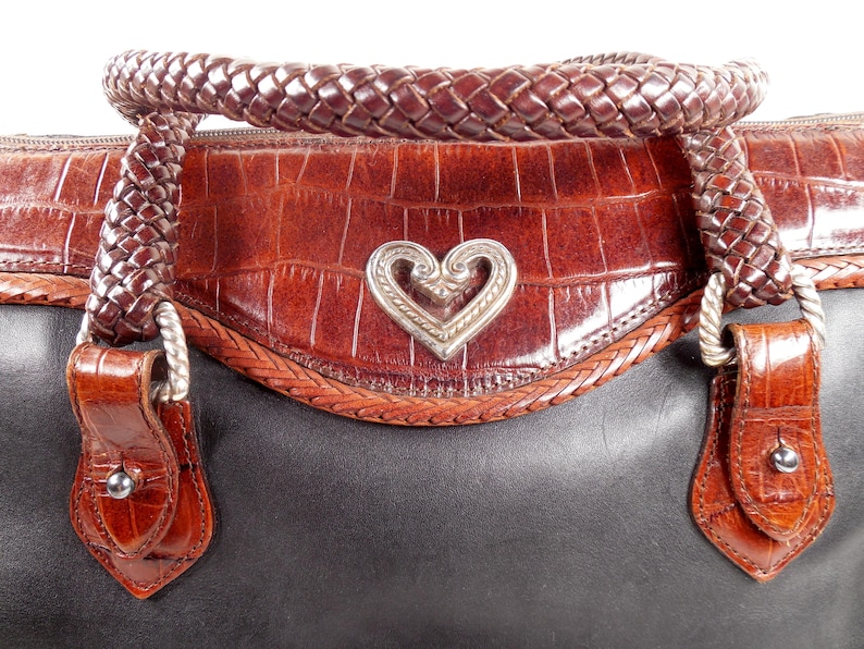 May include: A two-tone handbag featuring a smooth black leather base and a brown crocodile-embossed leather upper section. The bag includes a braided brown leather handle and a central silver-toned metal heart emblem with intricate scrollwork.