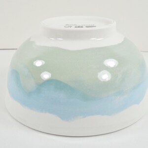 May include: A white ceramic bowl with a rounded shape. The bowl is painted with a watercolour design in shades of blue and green, creating a layered effect. The bottom and top rim are white.