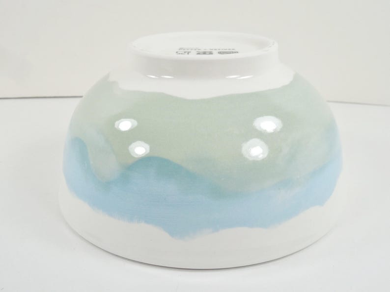 May include: A white ceramic bowl with a rounded shape. The bowl is painted with a watercolour design in shades of blue and green, creating a layered effect. The bottom and top rim are white.