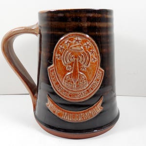 May include: A brown ceramic beer mug with a large handle. The mug features a raised emblem with text and a design of a radio tower and stars. The words "RAF Mildenhall" are on a banner below the emblem.