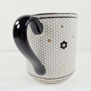 May include: White ceramic mug with a black handle. The mug features a honeycomb pattern with gold and black floral accents. The rim and base have a black and gold trim.
