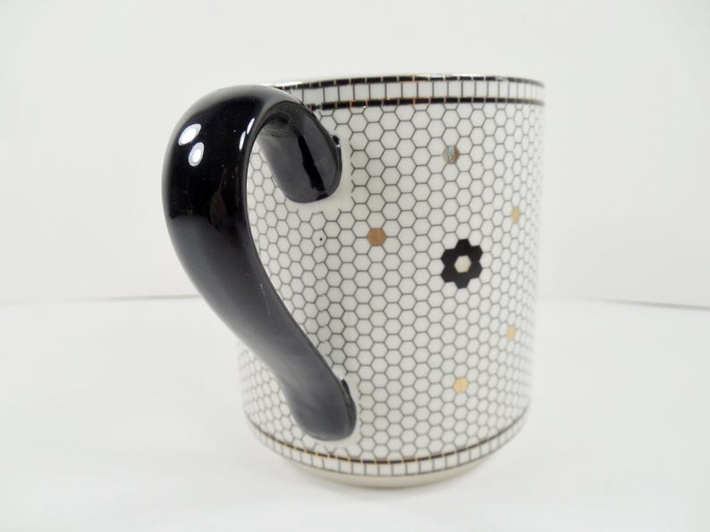 May include: White ceramic mug with a black handle. The mug features a honeycomb pattern with gold and black floral accents. The rim and base have a black and gold trim.