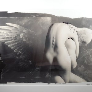 May include: A black and white photograph of a person with a white wing behind them. The person is nude and has their head covered with a white cloth. The person is hunched over and appears to be in pain.