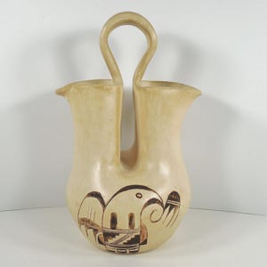 May include: A double-spouted ceramic vase with a unique handle. The vase is decorated with a brown and black geometric design.