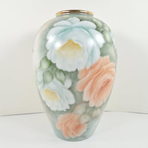 May include: A decorative vase with a painted floral design. The vase features large, detailed roses in shades of white, peach, and pale blue against a muted green background. The rim is accented with a gold band.