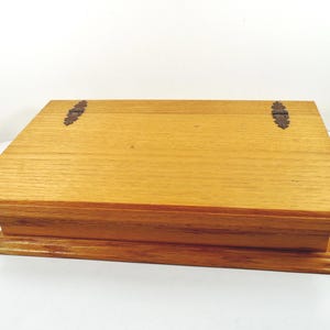 May include: A rectangular wooden box with a light brown finish. The box has a hinged lid with two decorative metal hinges. The box is made of wood and has a layered base.