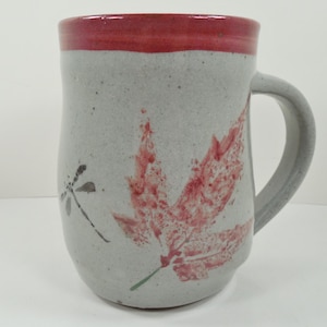 May include: A handmade ceramic mug with a gray speckled finish. The mug features a red maple leaf and dragonfly design. The rim is painted red, and it has a gray handle.