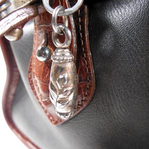 May include: A black handbag featuring a brown crocodile-embossed leather trim and a silver-toned metal zipper pull charm. The charm is shaped like a small, elongated capsule with an engraved leaf pattern and is attached to a silver metal ring.