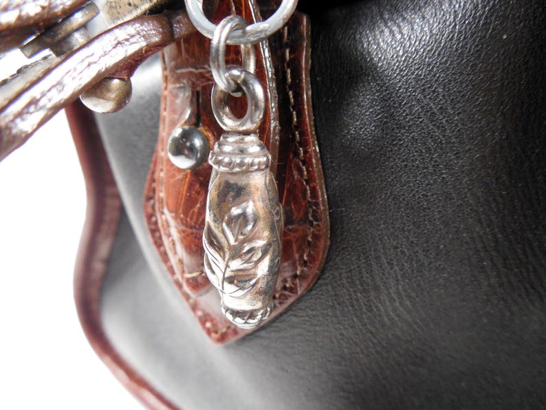 May include: A black handbag featuring a brown crocodile-embossed leather trim and a silver-toned metal zipper pull charm. The charm is shaped like a small, elongated capsule with an engraved leaf pattern and is attached to a silver metal ring.