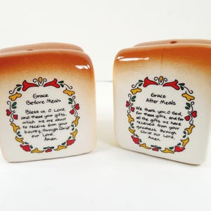 May include: A pair of ceramic salt and pepper shakers shaped like slices of bread. Each shaker has a white base with a floral design and text that reads "Grace Before Meals" and "Grace After Meals". The tops are brown.