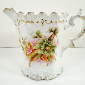 May include: White porcelain pitcher with a scalloped rim and handle, adorned with a floral design in shades of green, orange, and pink. The pitcher features gold accents and a decorative handle.