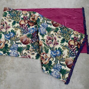 May include: A rectangular table runner featuring a cream-colored background with a dense floral and fruit pattern in shades of green, blue, purple, and brown. The reverse side is a solid magenta fabric with a textured finish and dark blue fringe.