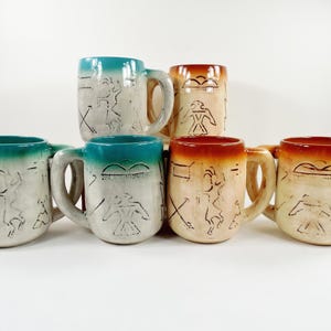 May include: A collection of five ceramic mugs featuring a light-colored base with dark etched petroglyph-style designs. Two mugs have a teal-colored rim and handle, while three mugs feature a warm orange-brown rim and handle design.