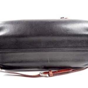 May include: A black leather handbag with a smooth finish and dark brown leather trim along the edges. The bag features a thin, adjustable brown leather shoulder strap with a silver-toned metal buckle and a small, dangling brown leather tassel.