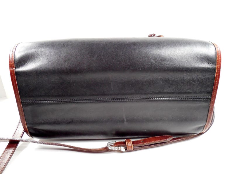 May include: A black leather handbag with a smooth finish and dark brown leather trim along the edges. The bag features a thin, adjustable brown leather shoulder strap with a silver-toned metal buckle and a small, dangling brown leather tassel.