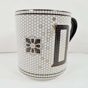 May include: White ceramic mug with a black handle and a mosaic tile design. The mug features a large black letter "D" outlined in gold, and a floral design. The background is a honeycomb pattern.
