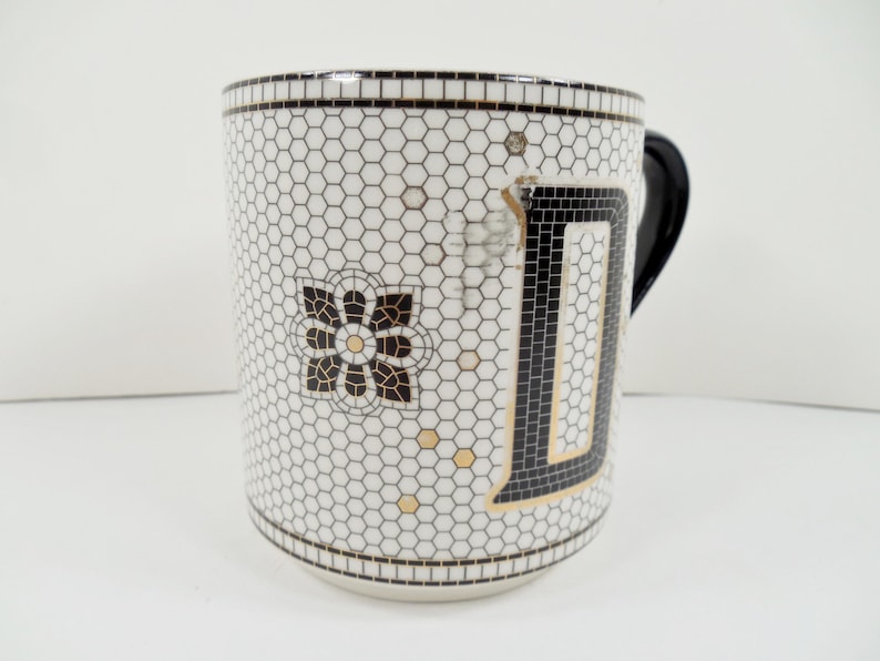 May include: White ceramic mug with a black handle and a mosaic tile design. The mug features a large black letter "D" outlined in gold, and a floral design. The background is a honeycomb pattern.