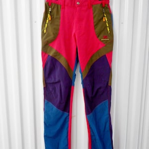 May include: Multi-colored outdoor pants with a vibrant design. The pants feature a red upper section, accented with olive green pockets and zippers. The lower sections are purple and blue. The brand name is visible on the right leg.