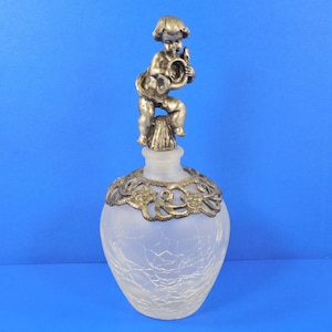May include: A frosted glass perfume bottle with a silver cherub figurine stopper. The cherub is playing a musical instrument and is sitting on a small base. The bottle has a decorative silver metal band around the neck.