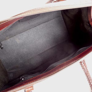 May include: An open brown leather handbag featuring a dark brown interior lining. The bag includes a zippered main compartment, a smaller interior zippered pocket, and braided leather handles attached to the top edges of the bag frame.
