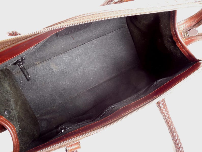 May include: An open brown leather handbag featuring a dark brown interior lining. The bag includes a zippered main compartment, a smaller interior zippered pocket, and braided leather handles attached to the top edges of the bag frame.