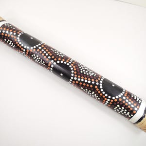 May include: A cylindrical musical instrument, a didgeridoo, with a black body adorned with white, orange, and brown dot patterns. The ends are wrapped with natural-colored woven material and white bands.