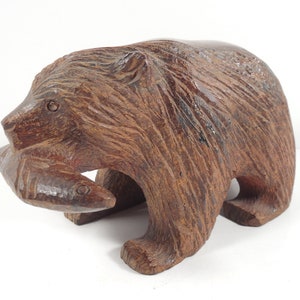 May include: A hand-carved wooden bear sculpture holding two fish in its mouth. The bear is brown and has a detailed, textured surface.