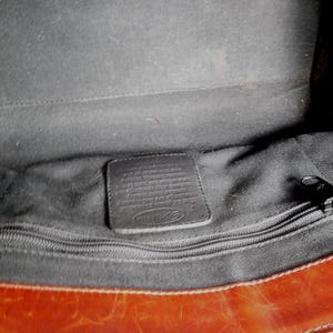 May include: The interior of a brown leather bag featuring a black fabric lining, a horizontal zippered pocket with a black metal pull, and a rectangular leather patch embossed with the Fossil brand logo centered above the zipper compartment.