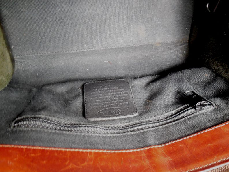 May include: The interior of a brown leather bag featuring a black fabric lining, a horizontal zippered pocket with a black metal pull, and a rectangular leather patch embossed with the Fossil brand logo centered above the zipper compartment.