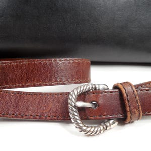May include: A brown leather strap with a silver-toned metal buckle featuring a twisted rope texture. The strap is positioned in front of a smooth black leather surface. The buckle has a prong and a single leather loop to secure the strap end.