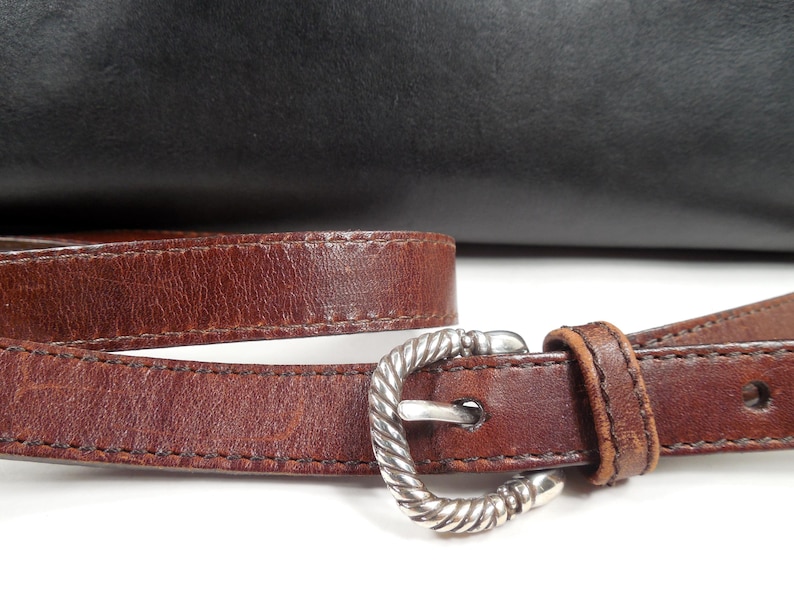 May include: A brown leather strap with a silver-toned metal buckle featuring a twisted rope texture. The strap is positioned in front of a smooth black leather surface. The buckle has a prong and a single leather loop to secure the strap end.