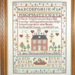 May include: Framed cross-stitch sampler with a house, trees, and floral motifs. The sampler includes the alphabet, numbers, and text that reads "In March + in April from morning to night..." The frame is a light brown color.