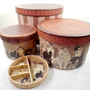 May include: A set of four decorative round boxes with lids. The boxes are various sizes and feature a cat-themed design. The lids are brown with paw print accents. The sides of the boxes are decorated with images of cats.