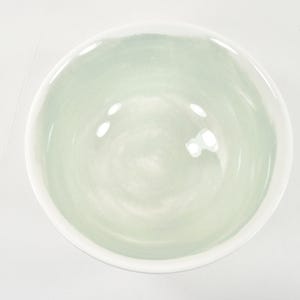 May include: A round ceramic bowl with a white rim and a pale green interior. The bowl has a smooth, glossy finish and is empty. The bowl is centered on a white surface.