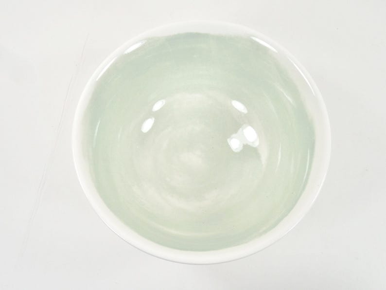 May include: A round ceramic bowl with a white rim and a pale green interior. The bowl has a smooth, glossy finish and is empty. The bowl is centered on a white surface.