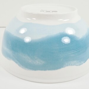 May include: A ceramic bowl with a white base and a gradient of blue hues. The bowl's design features a watercolour-like effect, transitioning from white to shades of light and medium blue. The bowl is likely used for serving food.