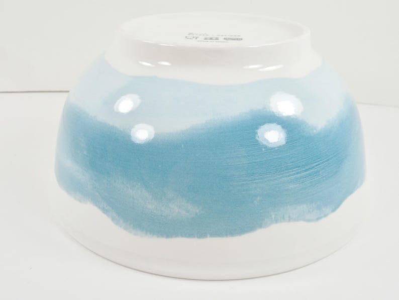 May include: A ceramic bowl with a white base and a gradient of blue hues. The bowl's design features a watercolour-like effect, transitioning from white to shades of light and medium blue. The bowl is likely used for serving food.